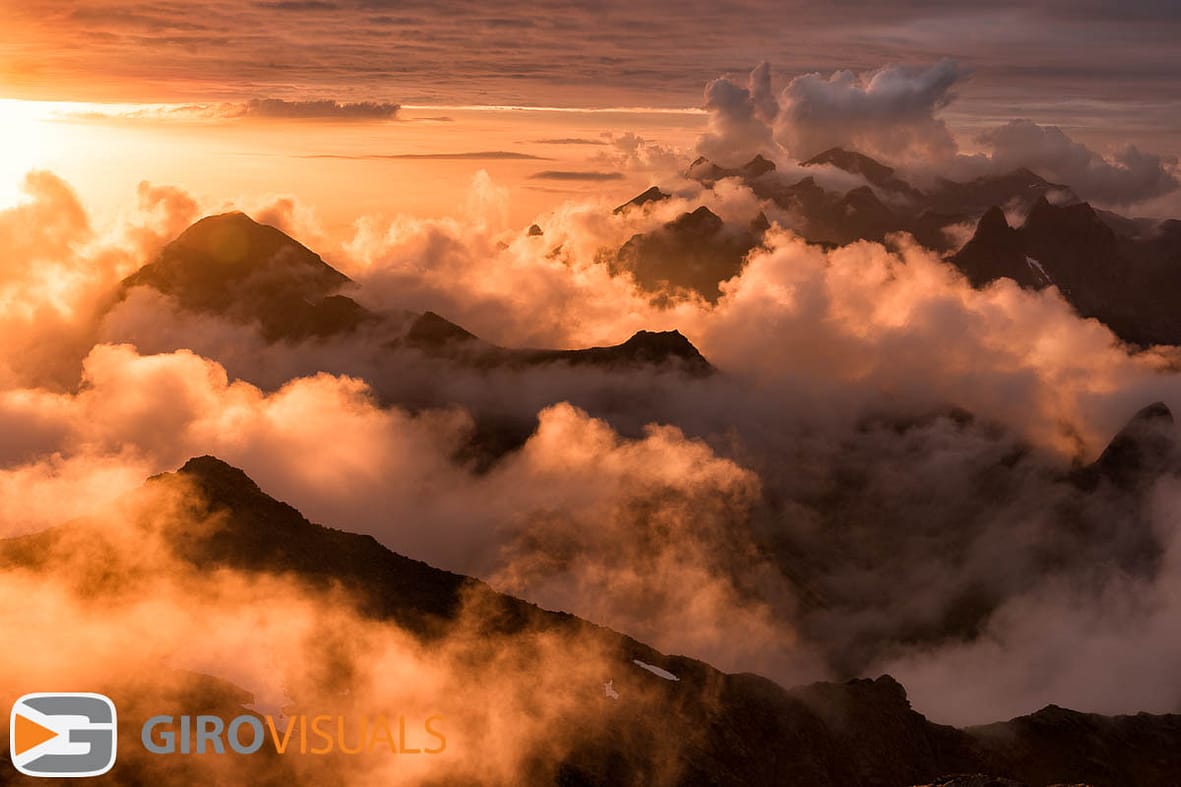 The midnight sun peaks below the cloud cover breaking thru the fog to light the numerous peaks that line the west coast of Lofoten Island, Norway